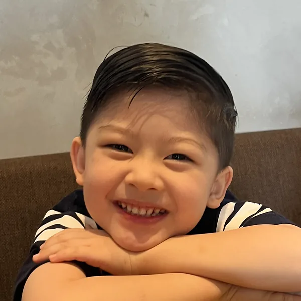 Henzel, 4 years old, resting his folded arms on a table and grinning at the camera.